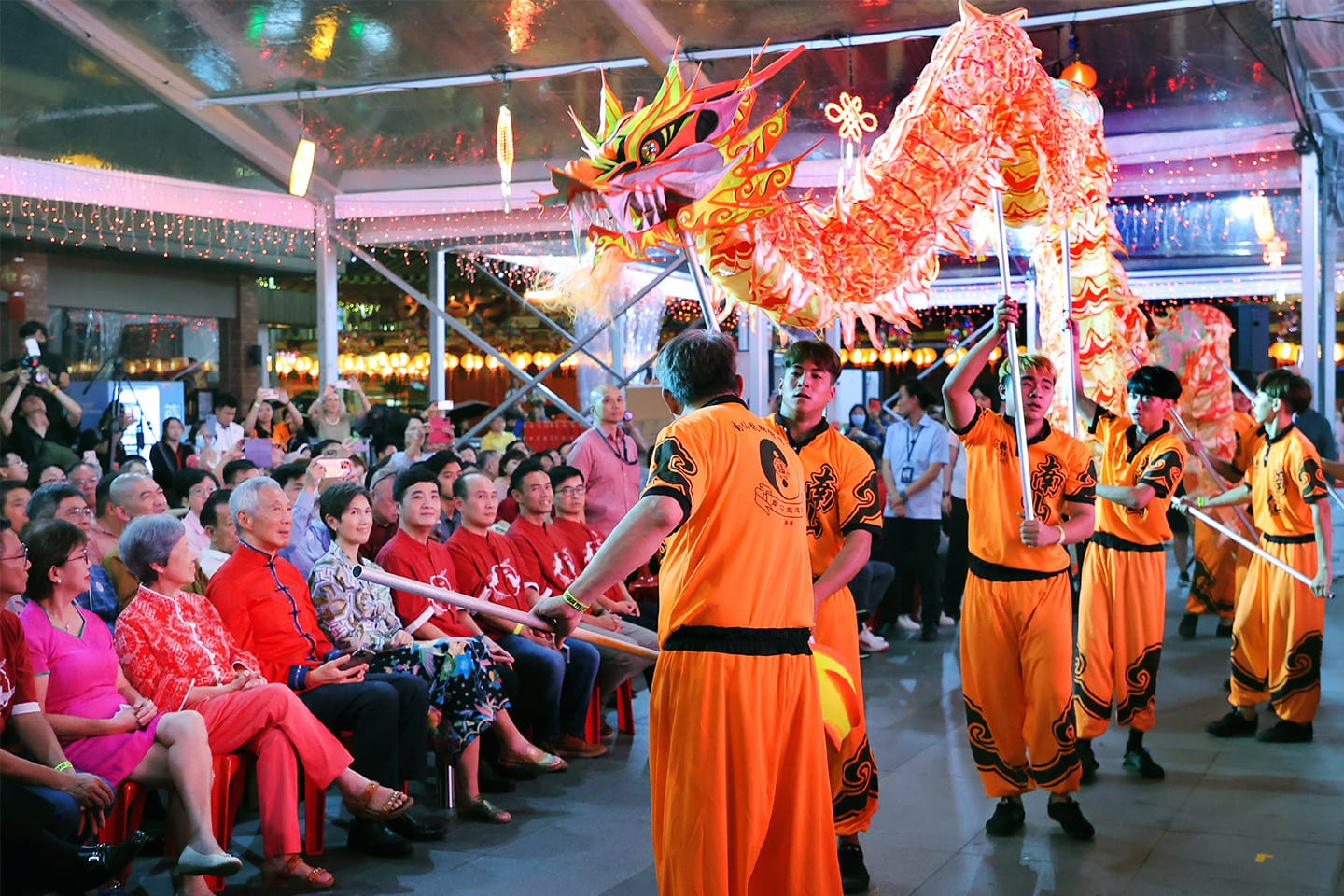Dragon dance performance with orange-clad performers and seated audience.